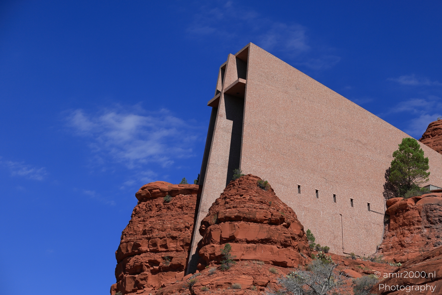 Chapel perched beside red rock formations, wide sky above and scattered desert shrubs below.