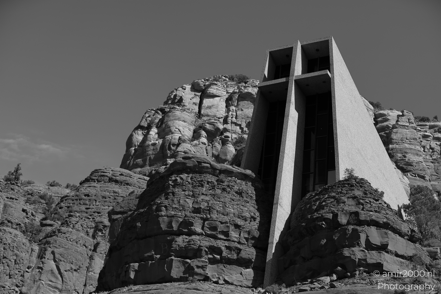 Black and white chapel framed by layered cliffs, strong contrast and bright sky highlights.
