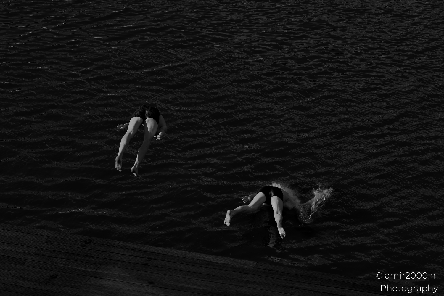 Two friends at the canal, one surfacing and one mid air, twin splashes forming in Amsterdam