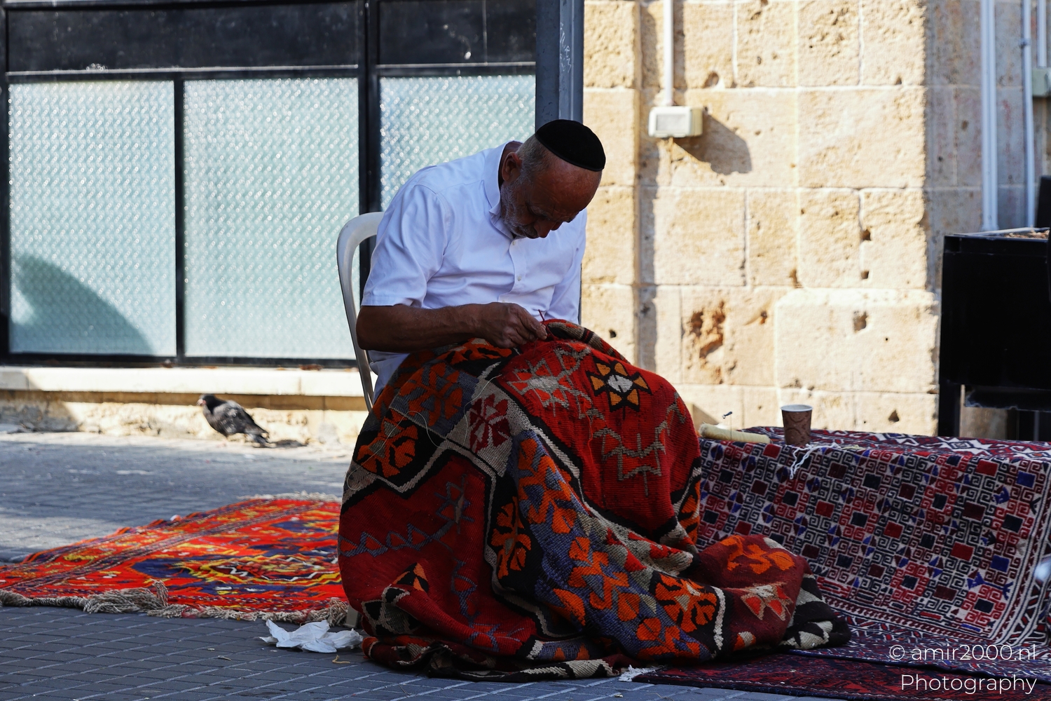 Craftsman seated outdoors pulling thread through a red patterned rug