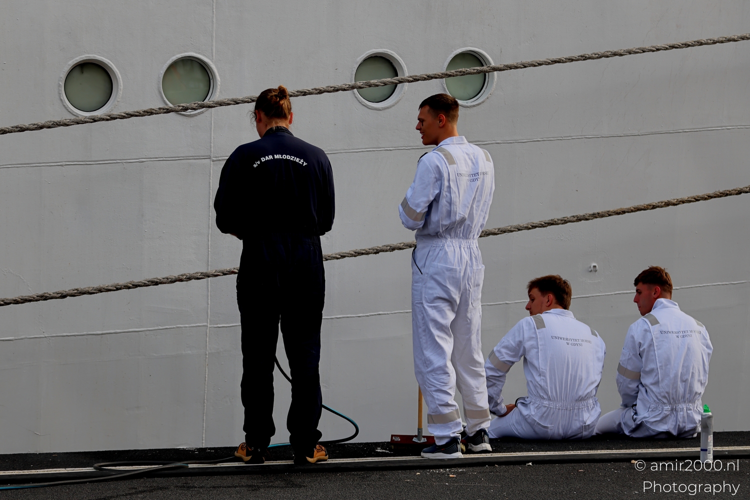 Ship crew in white coveralls beside portholes in Amsterdam harbor, soft overcast light, ropes crossing diagonally.