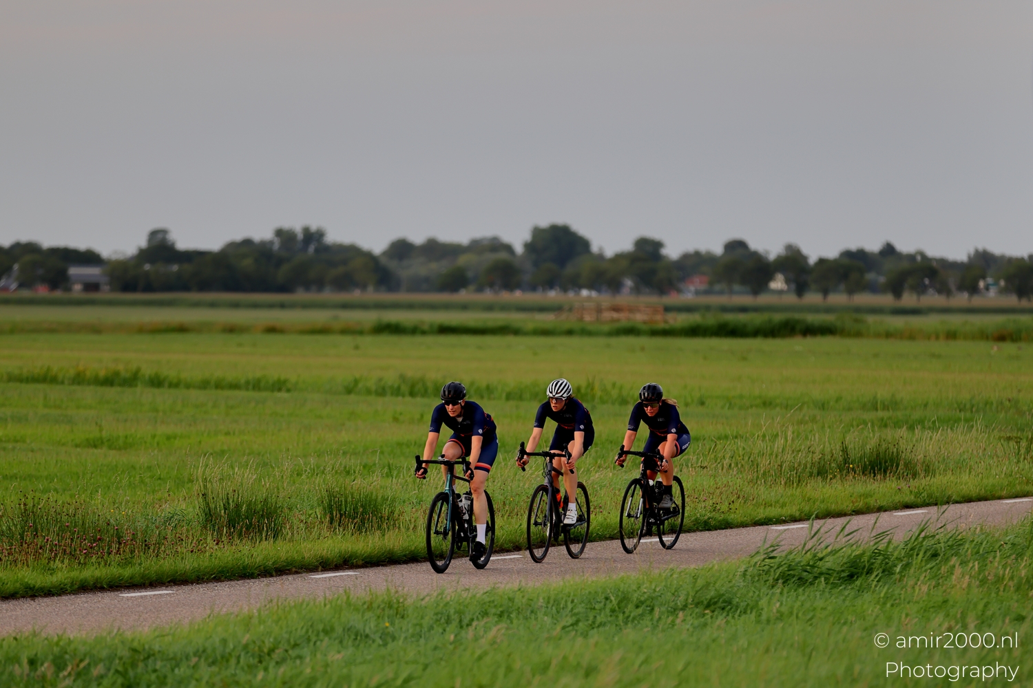 Three cyclists on a quiet road in Landelijk Noord near Amsterdam in soft evening light, long view with low horizon