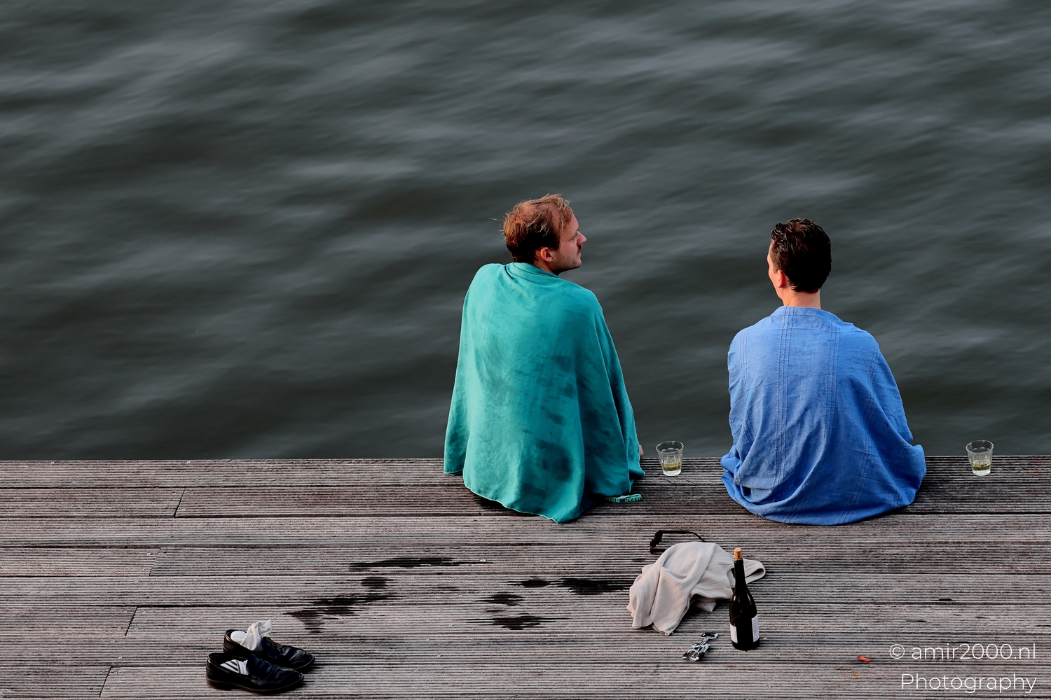 Two friends wrapped in towels on a wooden dock with drinks beside an Amsterdam canal at dusk