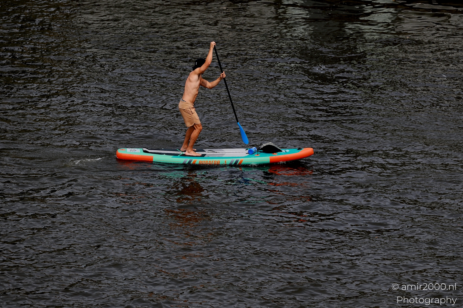Man paddleboarding on an Amsterdam canal with teal and orange board, overcast light, high angle to show ripples