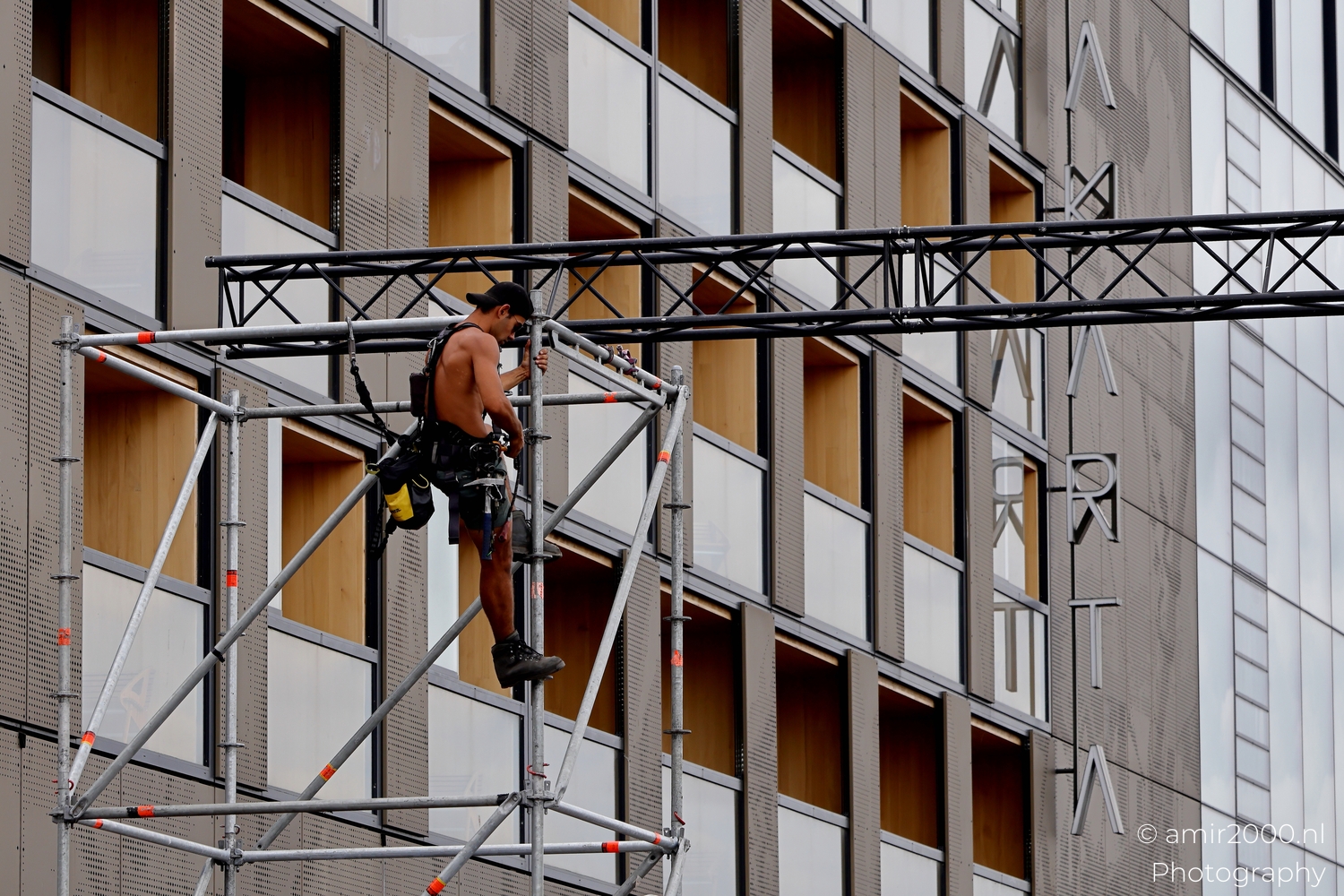 Two riggers on tall scaffold in Amsterdam with safety harnesses visible, vertical grid and grey sky holding tension.