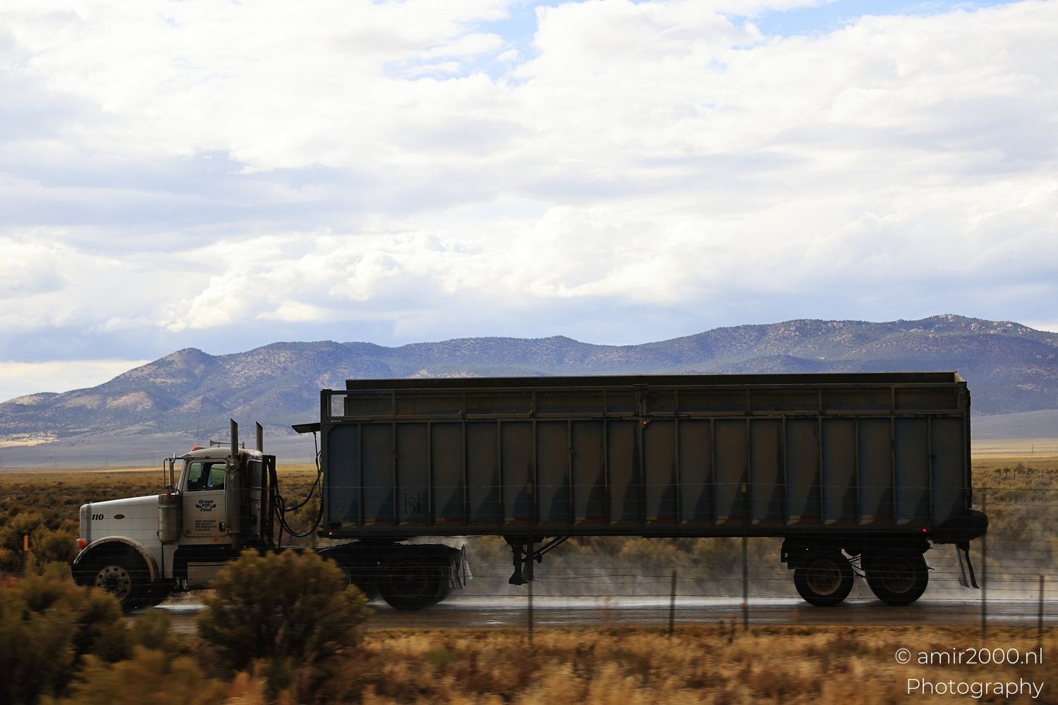White semi-truck transporting goods under cloudy skies along Interstate-15 near mountains and desert landscape.