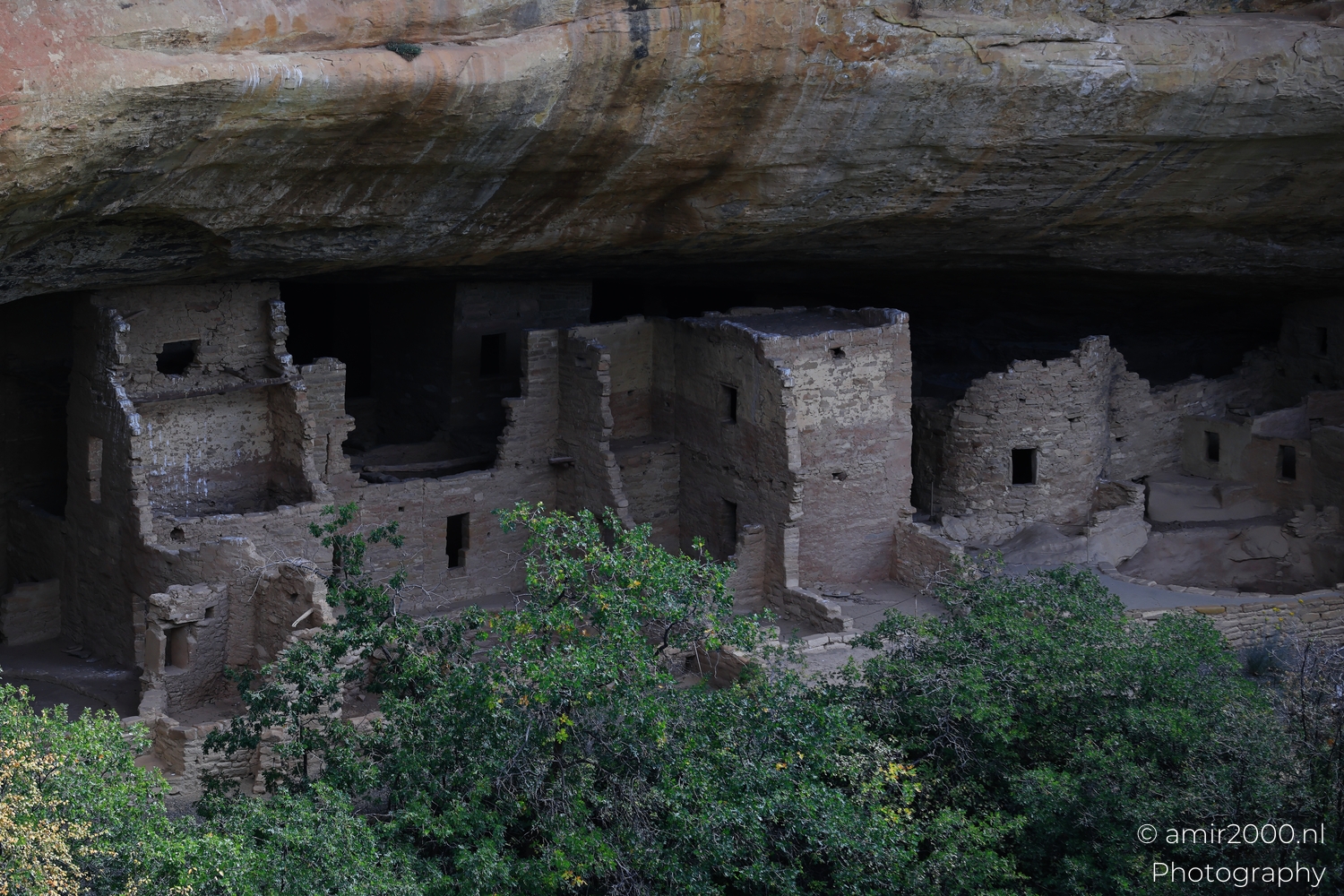 Cliff dwellings under massive sandstone overhang, distant viewpoint in shade, dark streaks on cliff face.