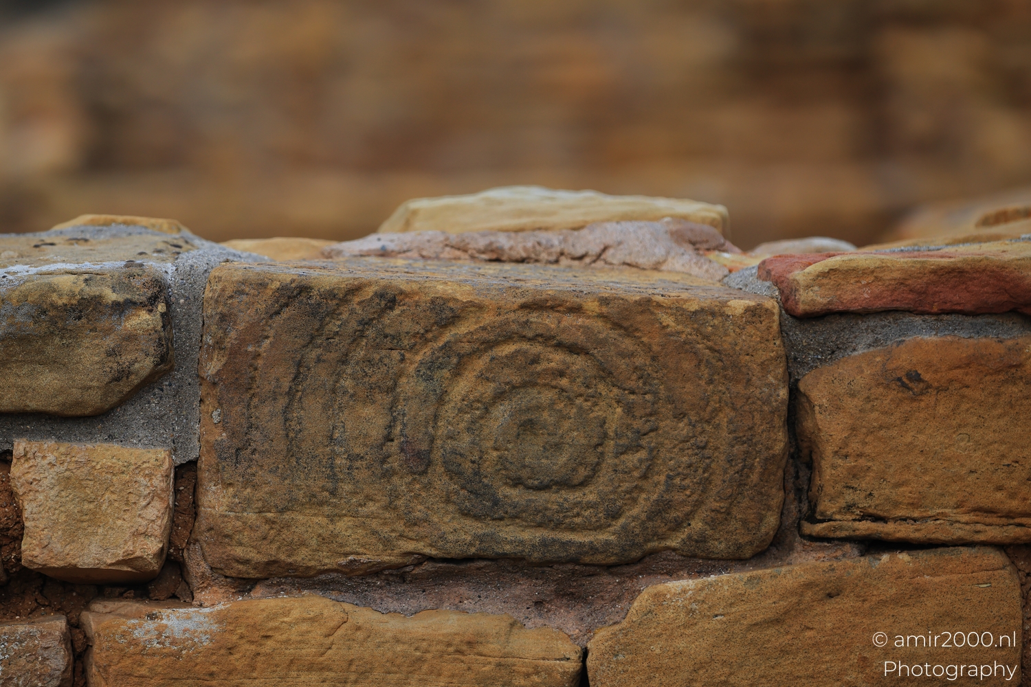 Spiral carving on weathered stone block, close view in soft light, sandy mortar seams.