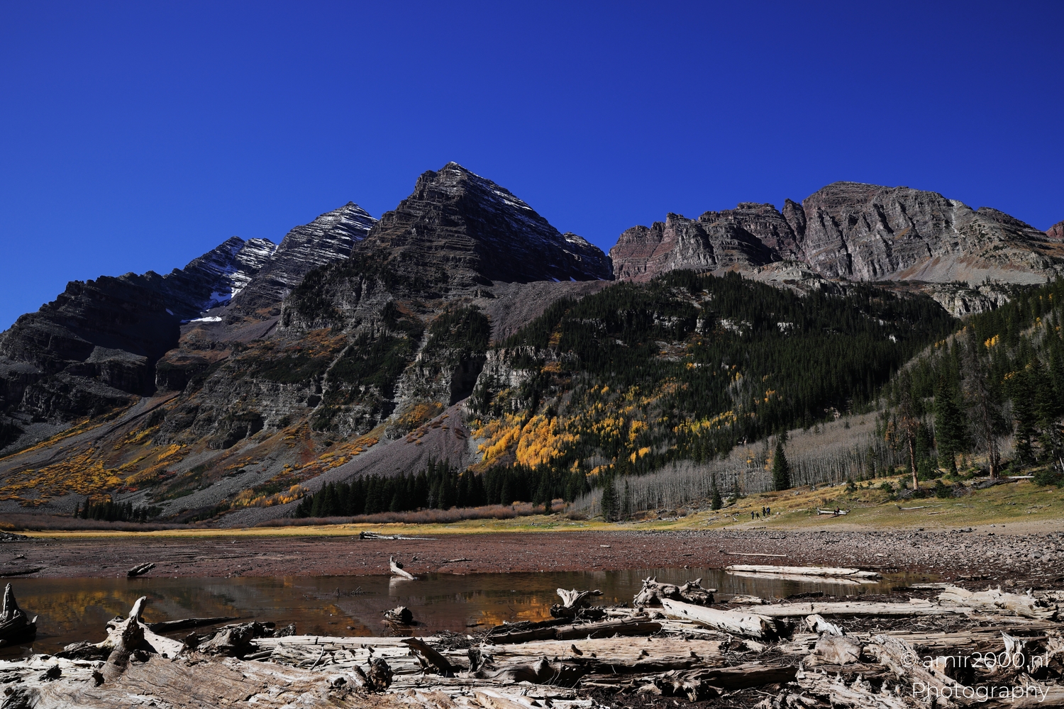 Wide valley scene with driftwood foreground, clear midday light, jagged peaks above shallow water.