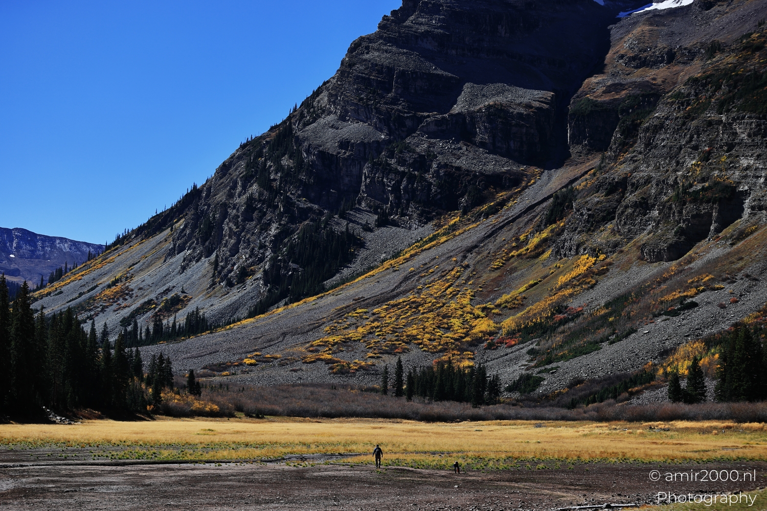 Long slope wall from meadow edge, bright daylight, lone hiker for scale near open grass.
