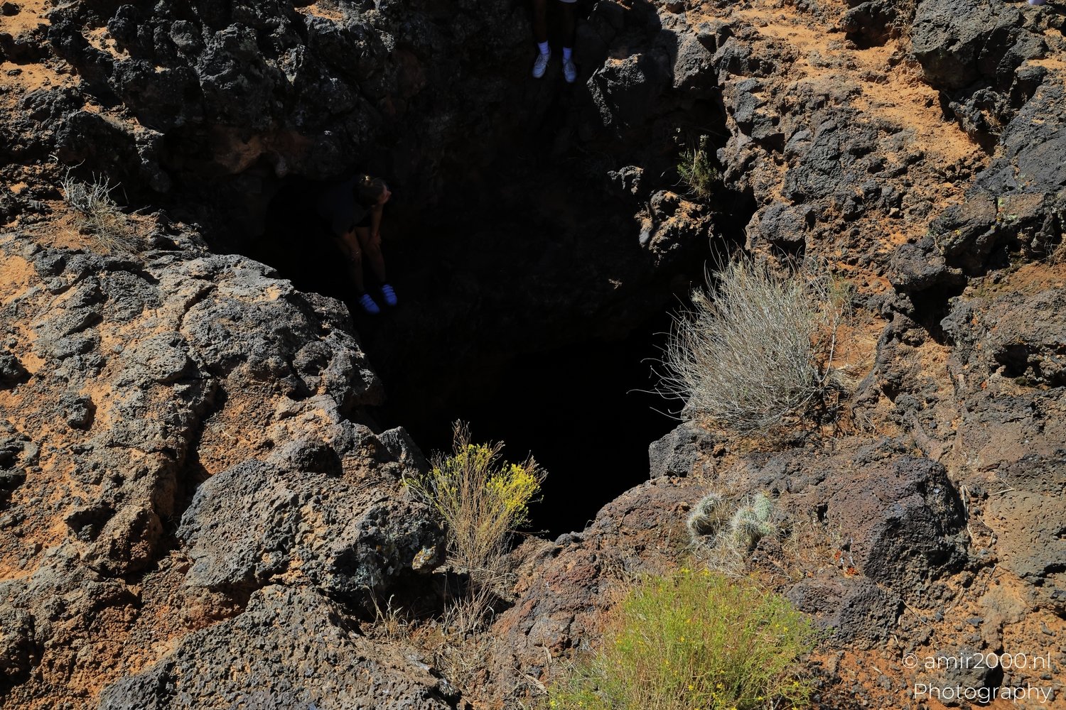 Person descending rocky desert cave with sparse vegetation and blue footwear visible.