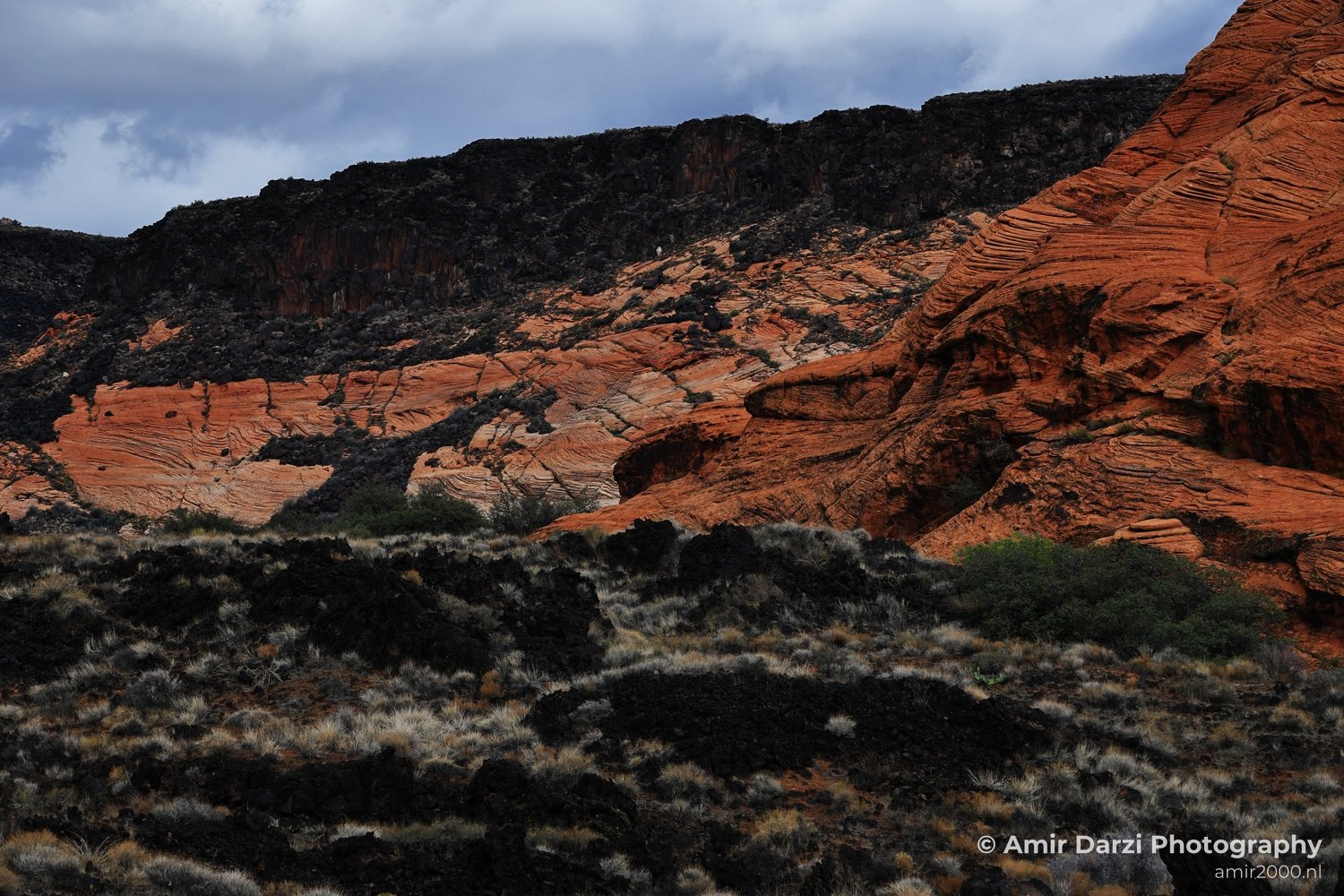 Layered red rock strata with sparse vegetation under a cloudy sky.