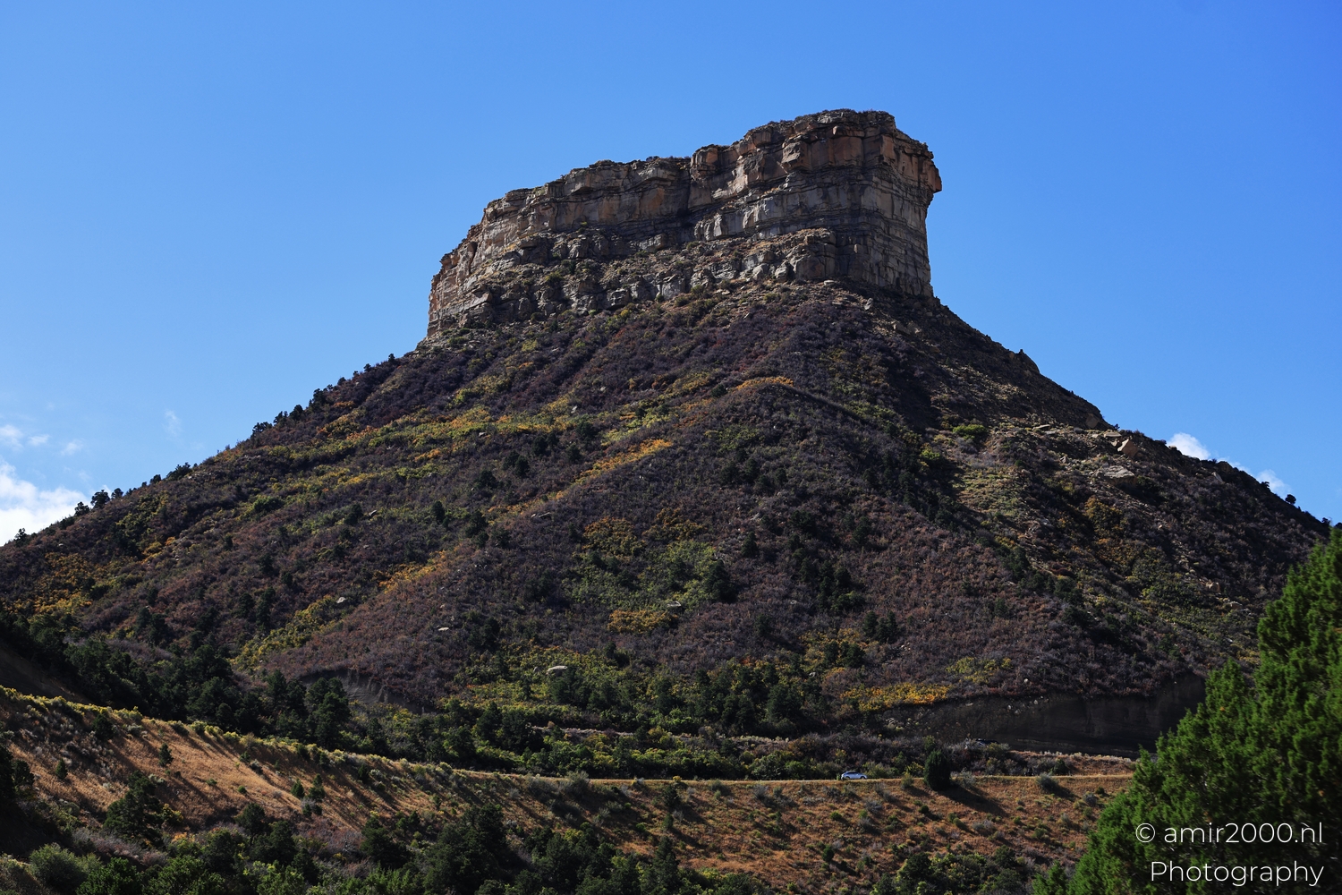Isolated mesa butte under clear blue sky, low angle view in bright sun, tiny car below.