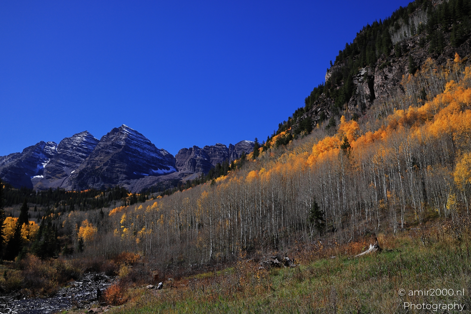 Mountain view with aspen hillside, cool daylight, winding stream and green foreground grasses.
