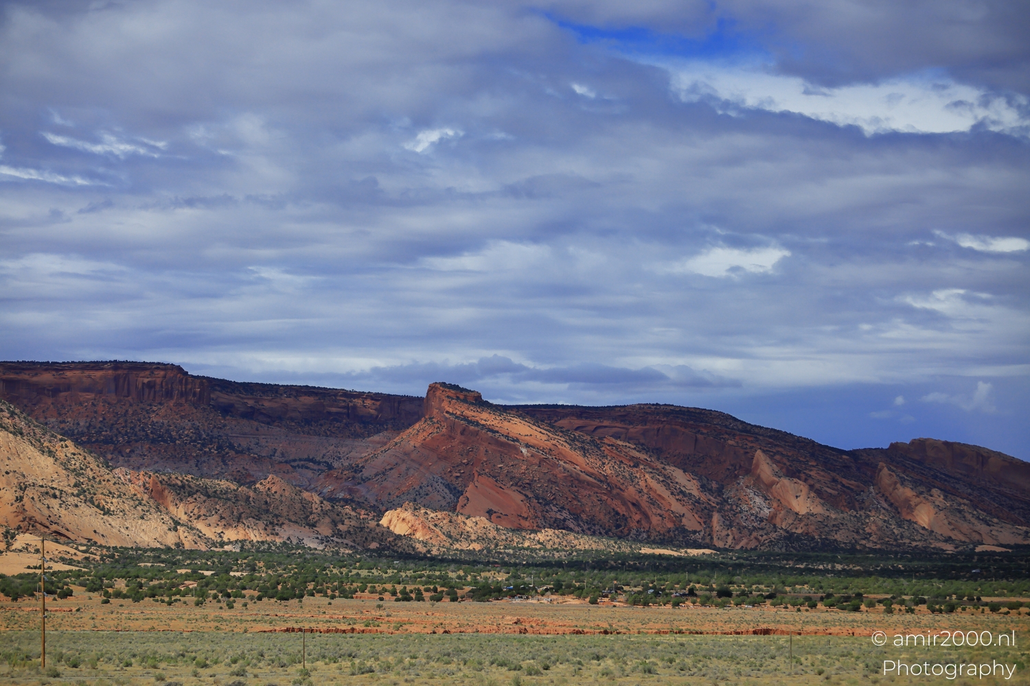 Red sandstone ridge from roadside view under layered clouds, with green trees across the valley.