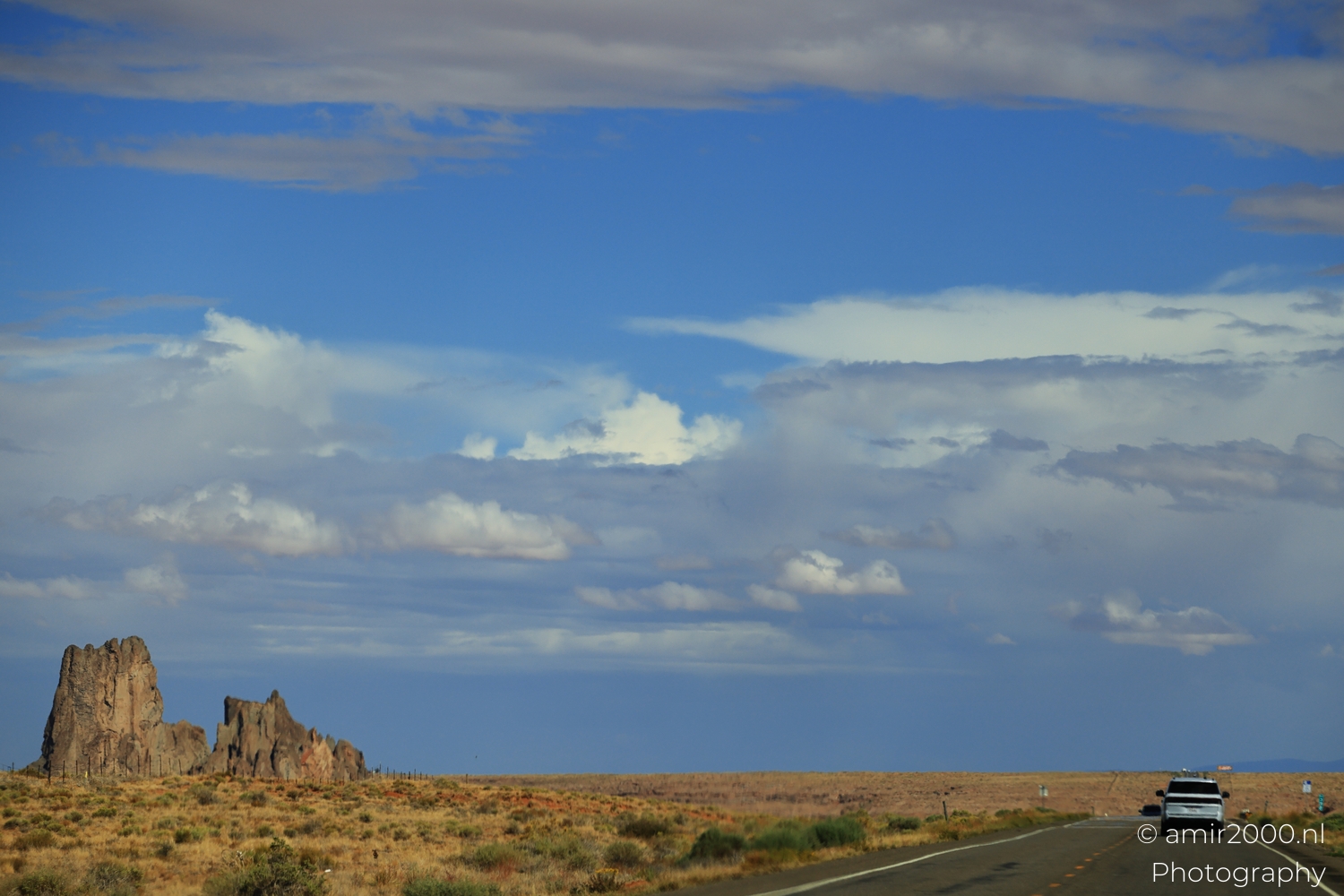 Highway curve from a moving car, with rock outcrops on the left and a vehicle ahead.