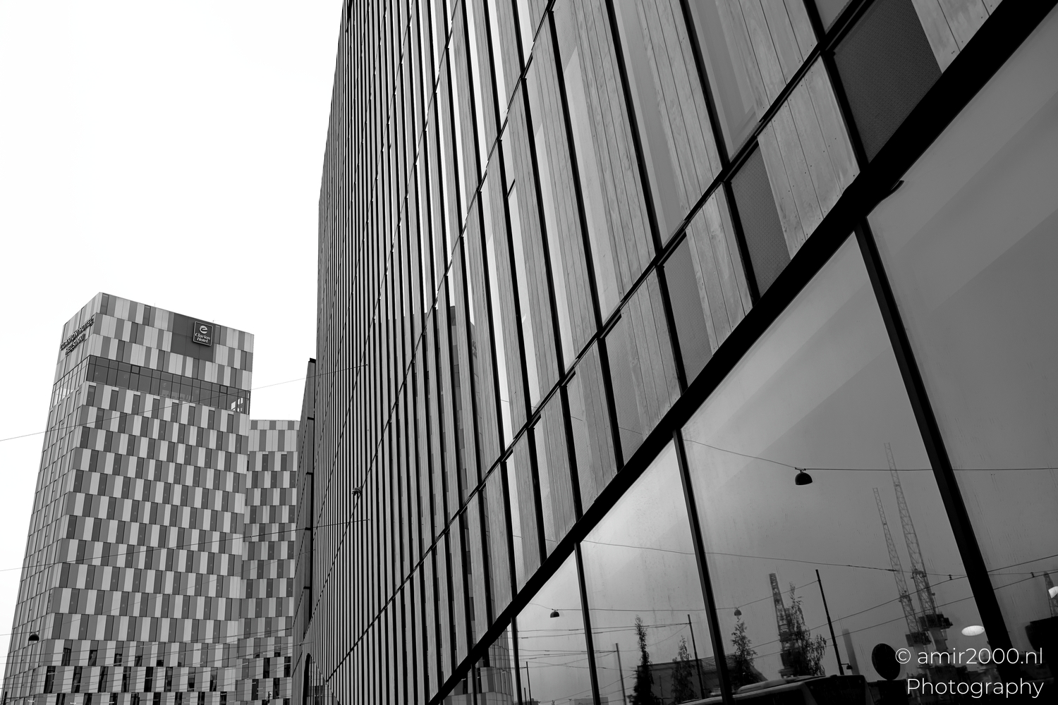 Side facade reflecting cranes street lamps and the hotel tower across a calm roadway on a grey day.
