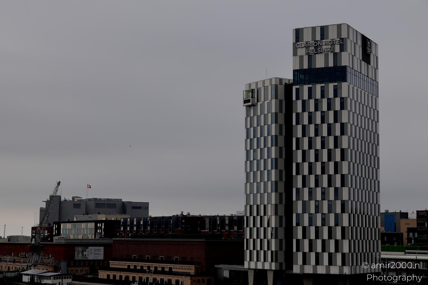 Distant view of Clarion Hotel Helsinki rising above harbor buildings beneath uniform grey clouds and cranes.