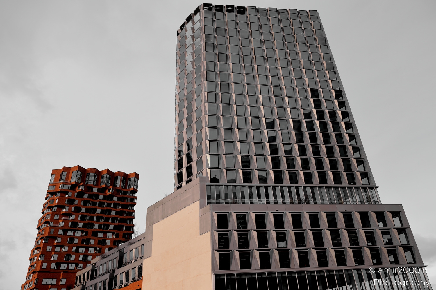 Angled glass tower with repeating facets and a brick neighbor in Amsterdam Zuidas under soft grey sky