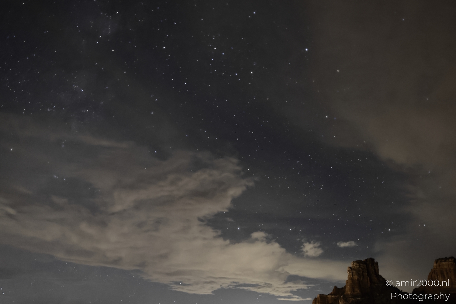 Cloud bands crossing starry night sky above desert ridge, wide view, subtle horizon light dome