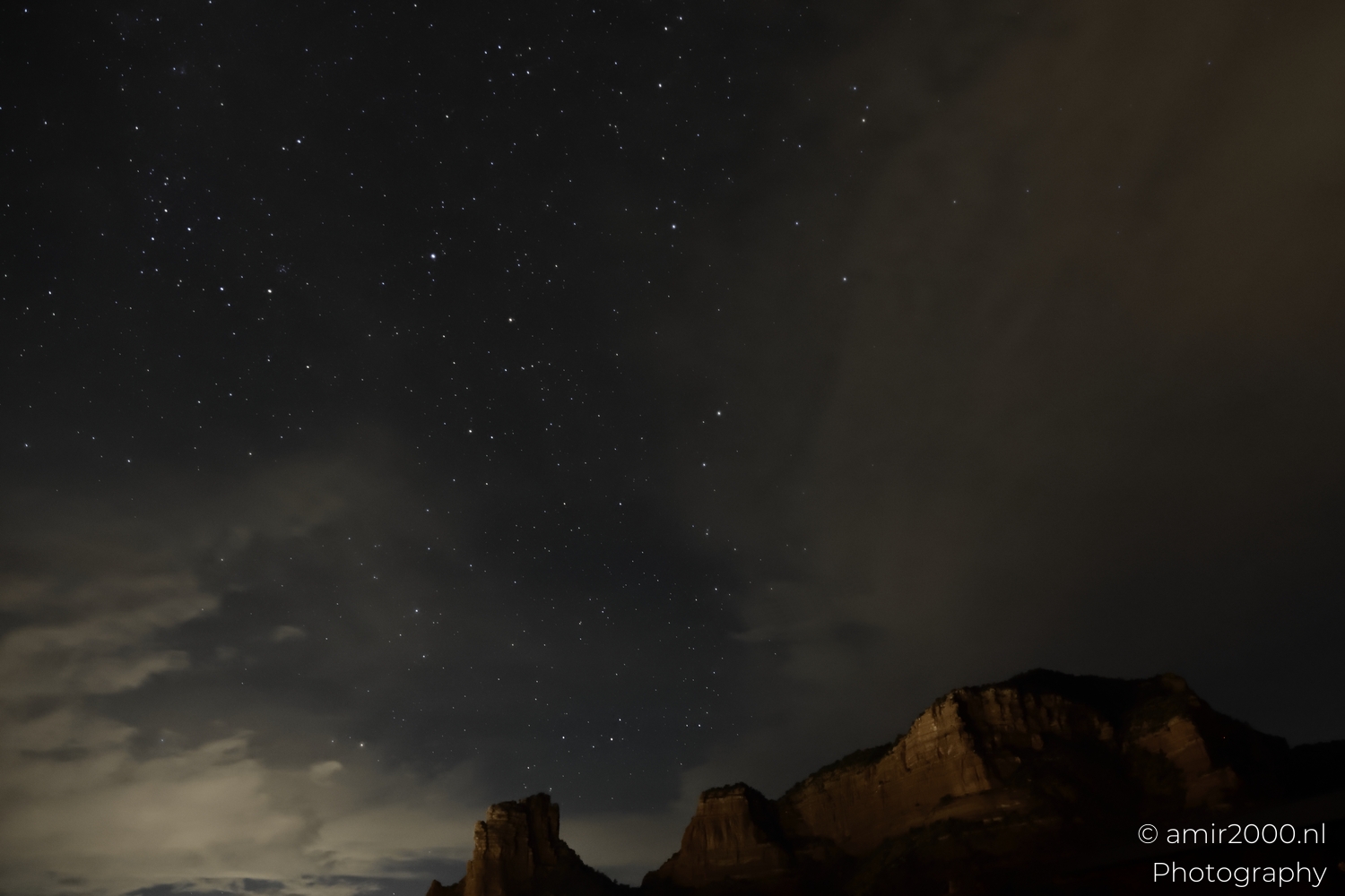 Starfield above sandstone outcrop at right, soft cloud haze, wide desert night composition