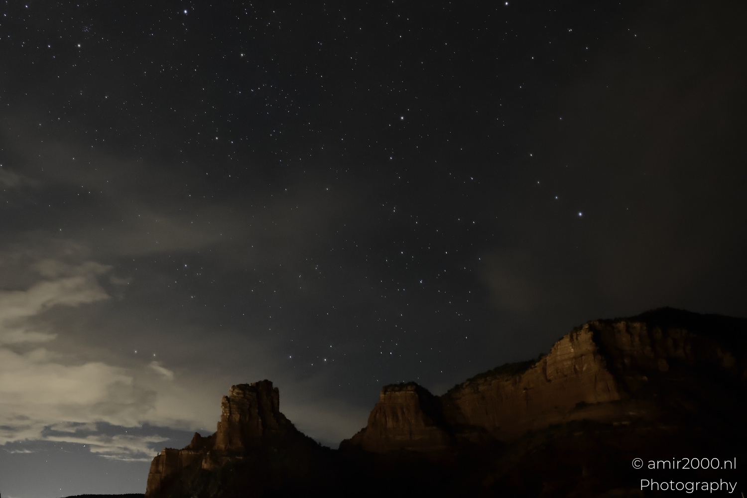 Rocky desert foreground under scattered clouds, stars in clear gaps, wide night landscape view
