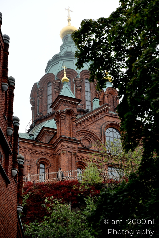 Red brick walls and domes of Uspenski Cathedral partially hidden by leafy trees on an overcast day.
