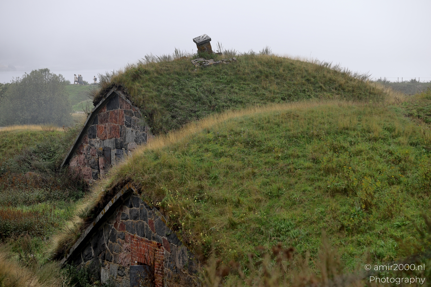 Rounded grass covered bunkers at Suomenlinna with stone entrances and distant figures in soft fog