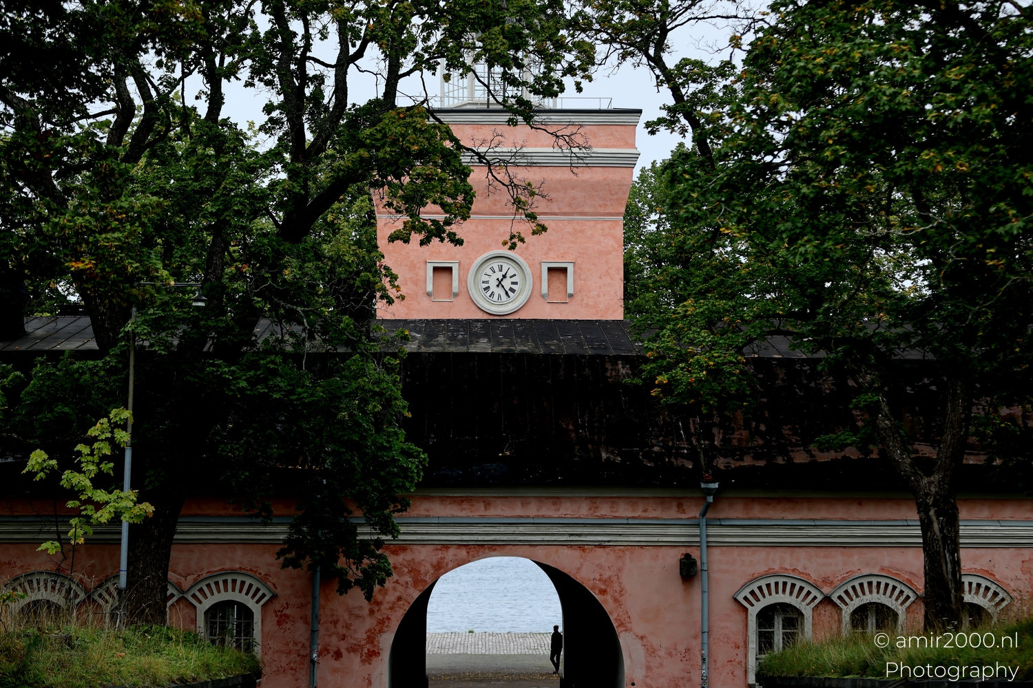 Pink clock tower at Suomenlinna framed by trees above an archway leading toward the sea on a misty day