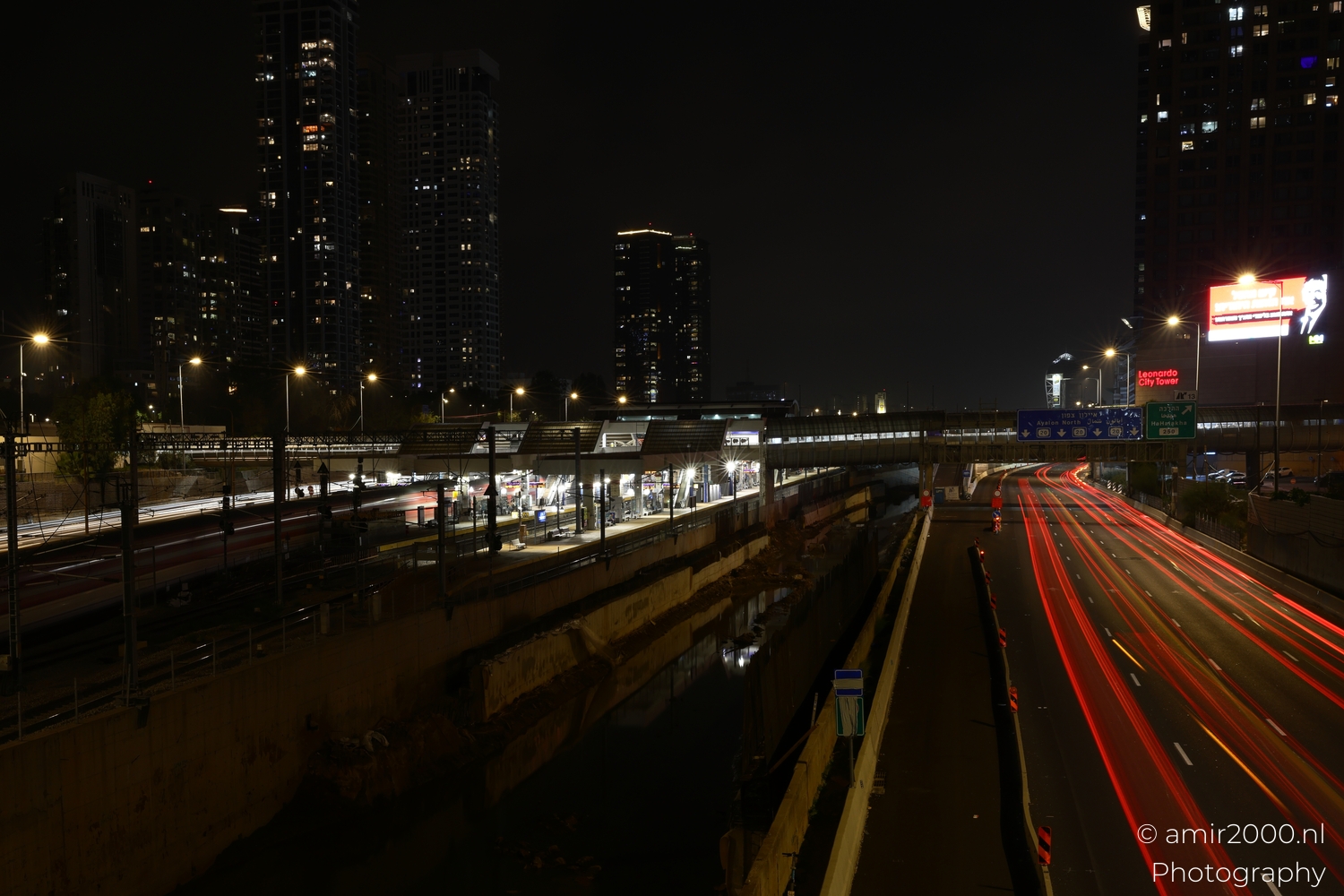 Rail platforms and road light trails split the scene, framed by tall residential towers at night.