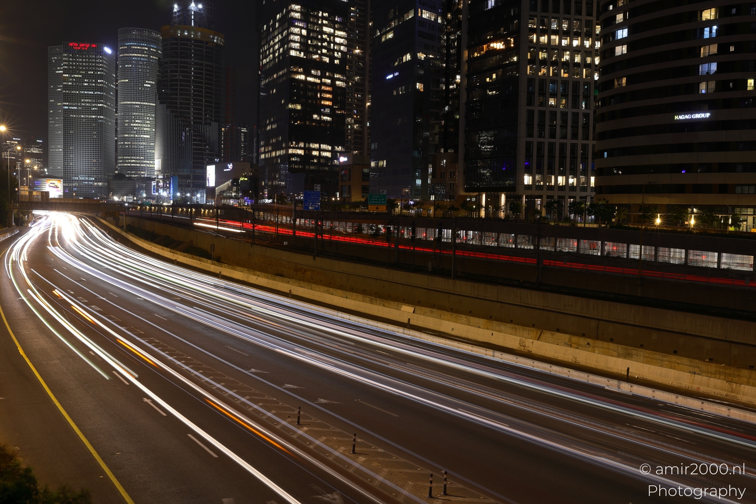 Broad sweep of traffic light trails with dense glass towers and a dark canal mirror below.