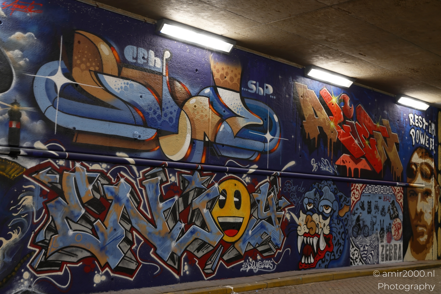 Colourful graffiti wall with bold lettering, smiling yellow face and cartoon like monster under strip lights at night