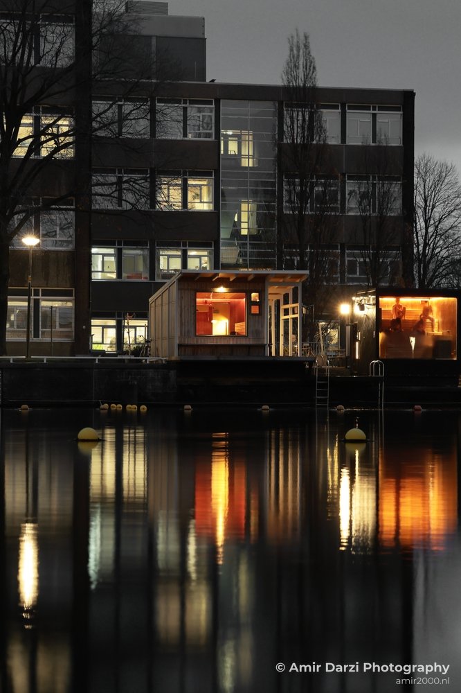 An illuminated sauna with reflective water and surrounding urban buildings.