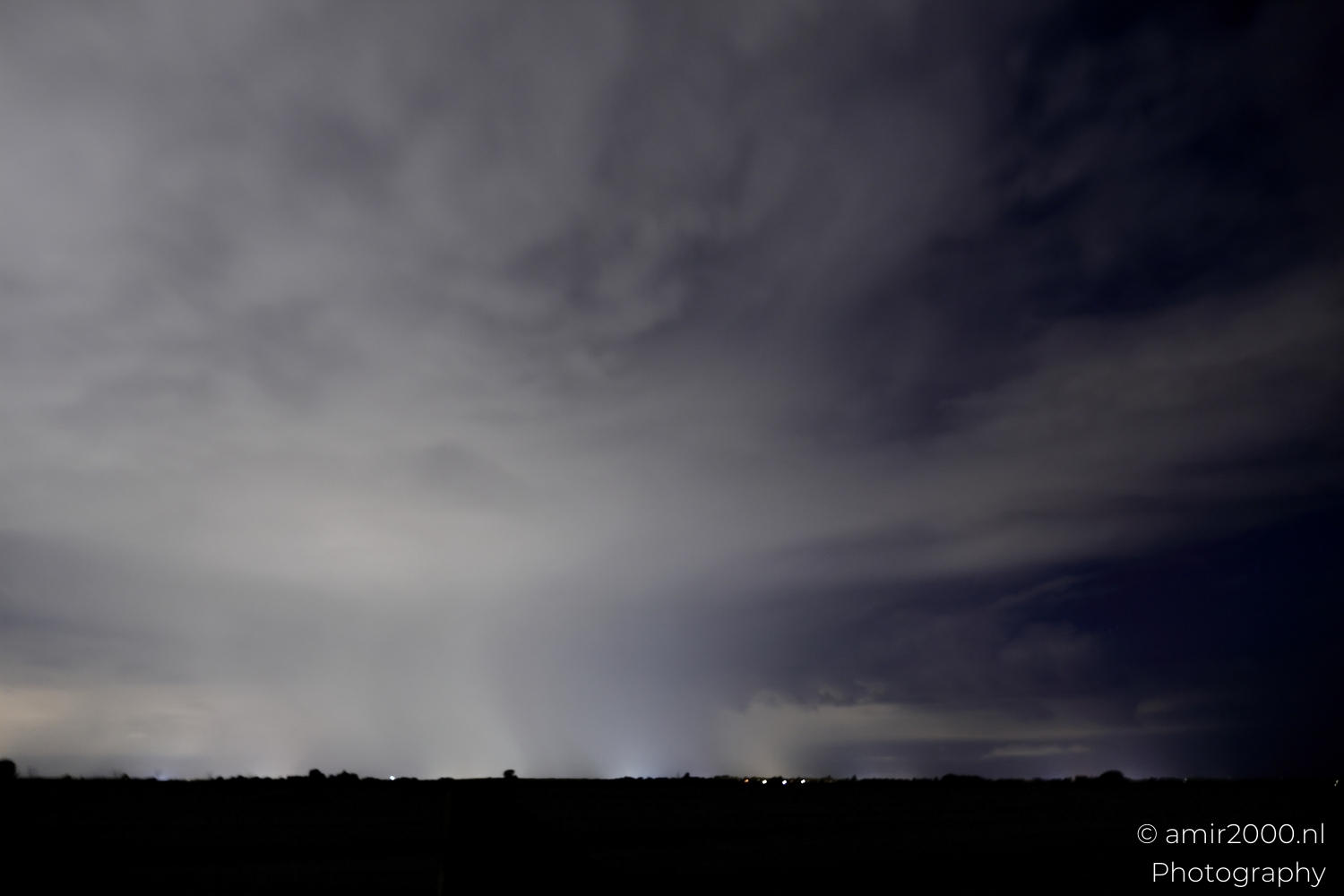 Bright rain shaft glowing beneath a thick cloud lid with soft urban glow along the base.