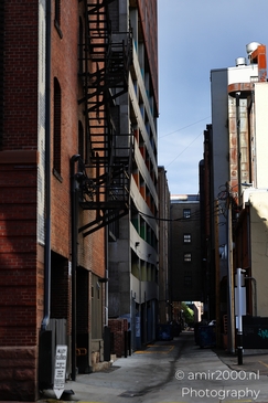 A narrow urban alleyway with red-brick industrial-style buildings and a fire escape. - image from year 2025 #023
