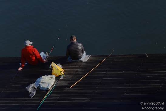 Two men fishing on a wooden pier in Amsterdam canal, in Amsterdam Netherlands. . - image from year 2025 #001