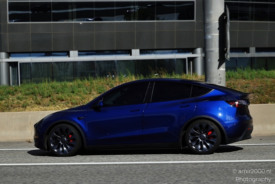 A vibrant blue Tesla Model Y is driving on a highway with an industrial building in the - image from year 2025 #013