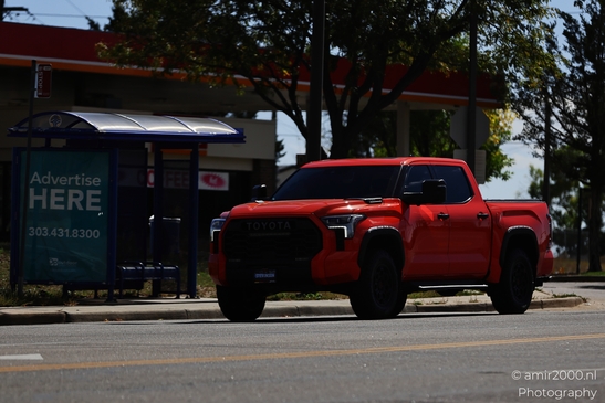 A vibrant red Toyota Tundra pickup parked beside a bus stop and gas station on the roadside. - image from year 2025 #002