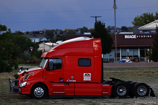 A large red semi-truck parked in a dirt lot with buildings and trees visible. - image from year 2025 #001