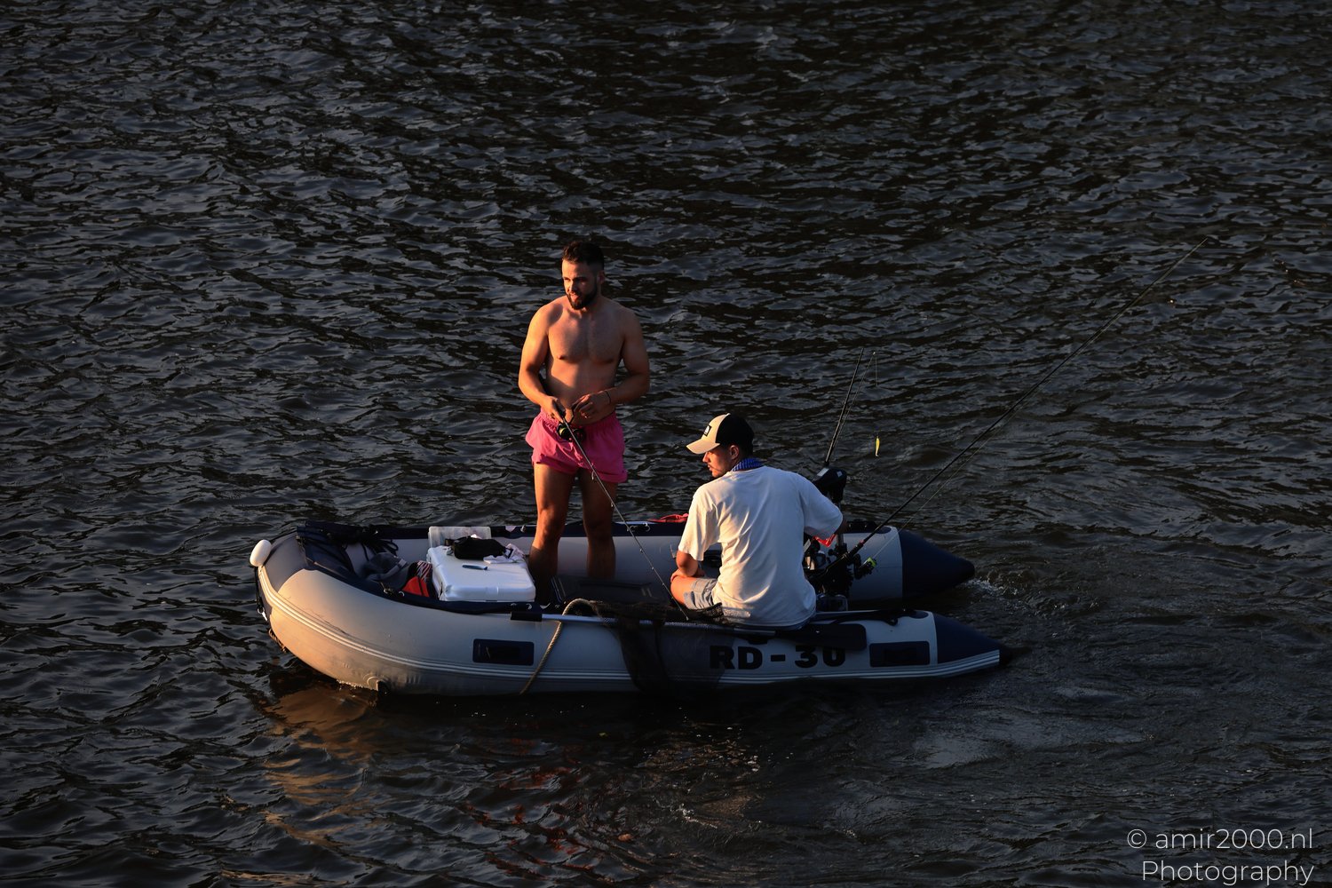 A shirtless man and another wearing a cap are fishing from an inflatable raft on a lake in Amsterdam, Netherlands.