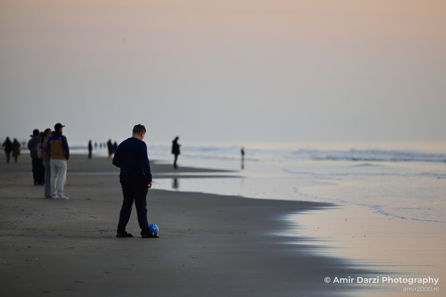 A solitary figure on a beach at sunset, kicking a soccer ball, with the horizon and distant figures.