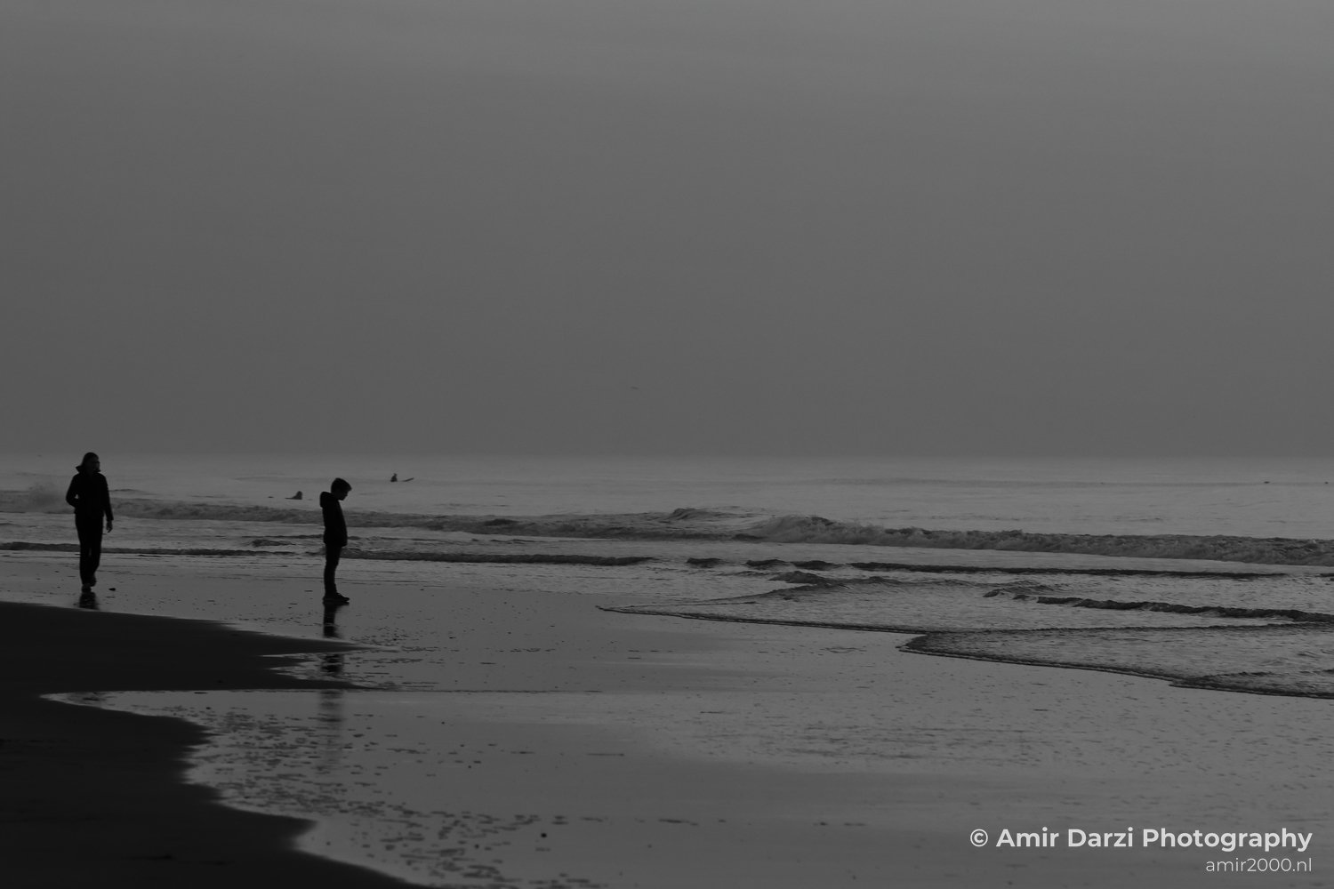 A beach scene at twilight, featuring two people walking and waves crashing on the shore.