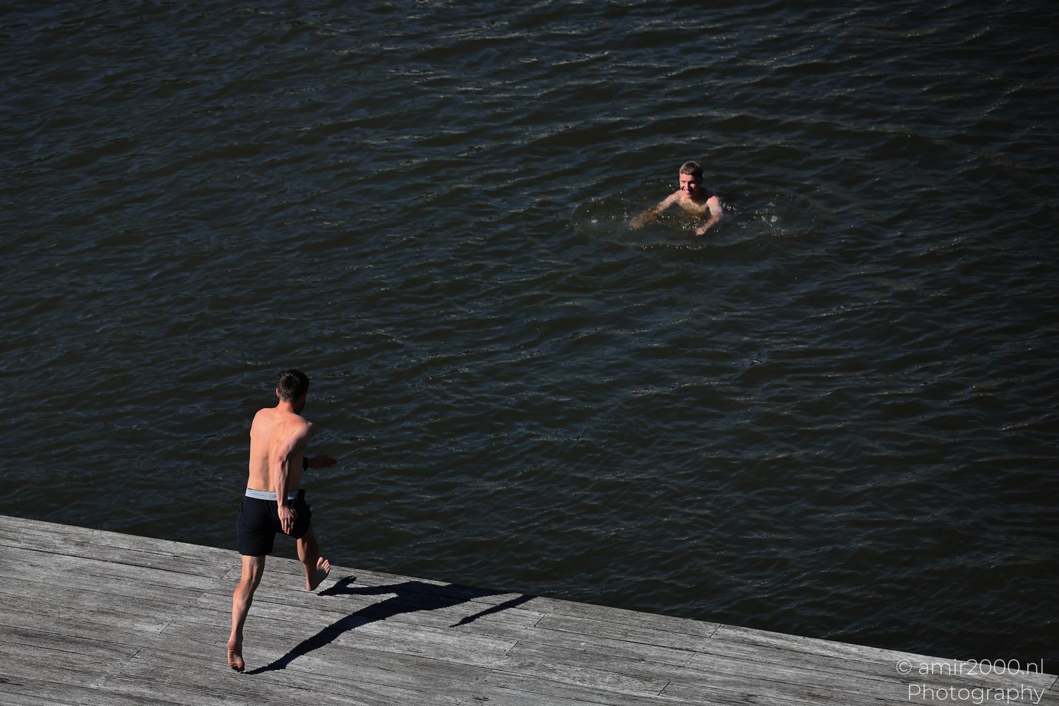 Jump Canal a swimmer in the distance and a diver at the edge of a wooden pier in water in Amsterdam, Netherlands.