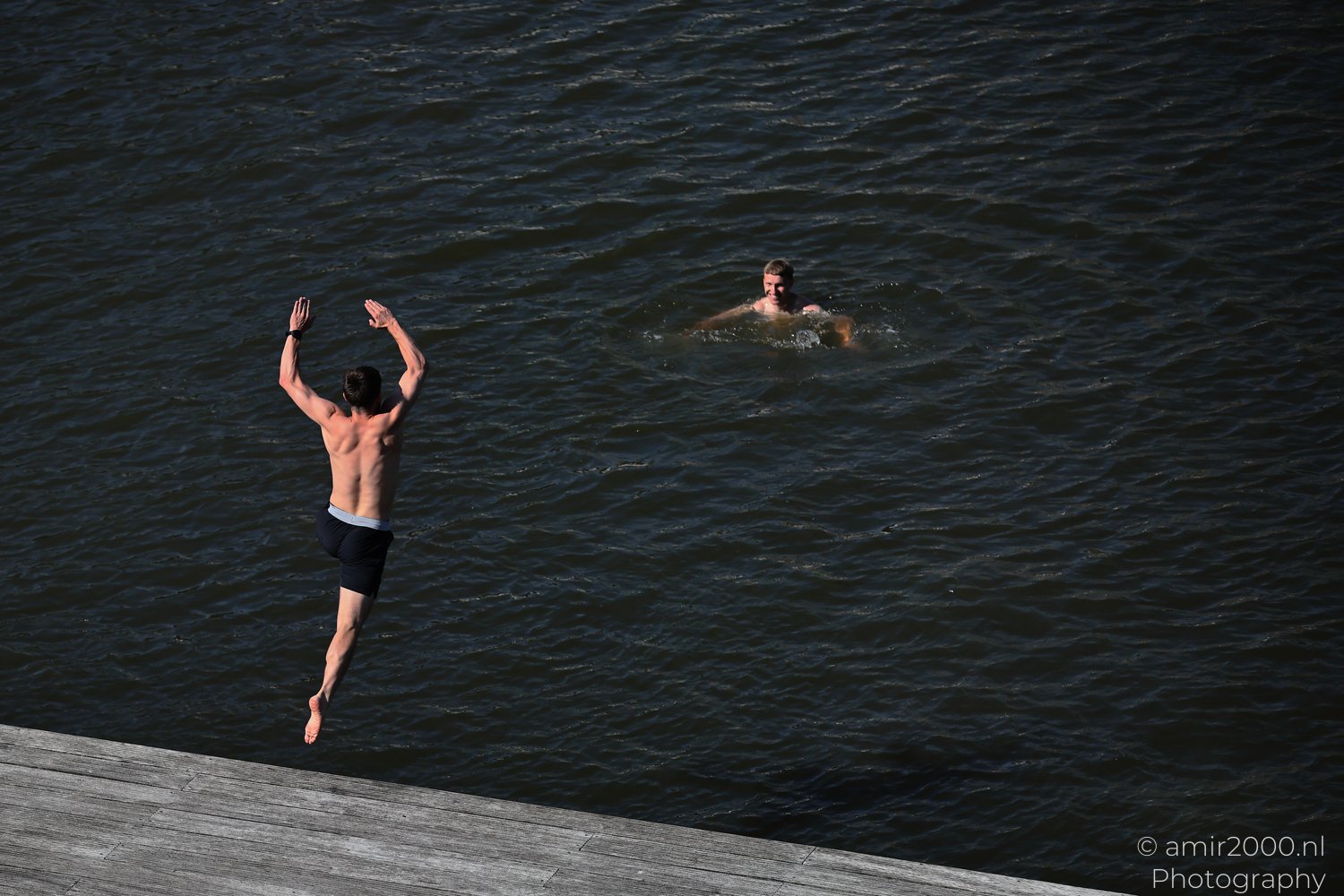 Jump Canal an individual is diving from a wooden pier into a water as another person swims in Amsterdam, Netherlands.