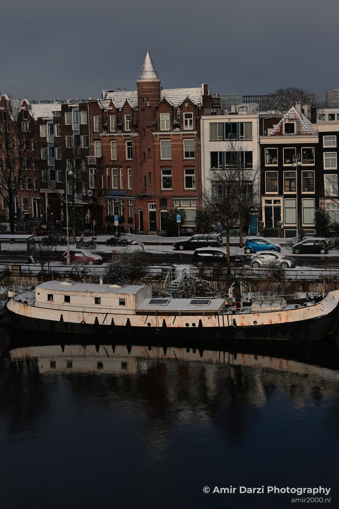 Winter scene of an old boat moored along Wittenburggracht Canal with snow covered buildings and parked cars.