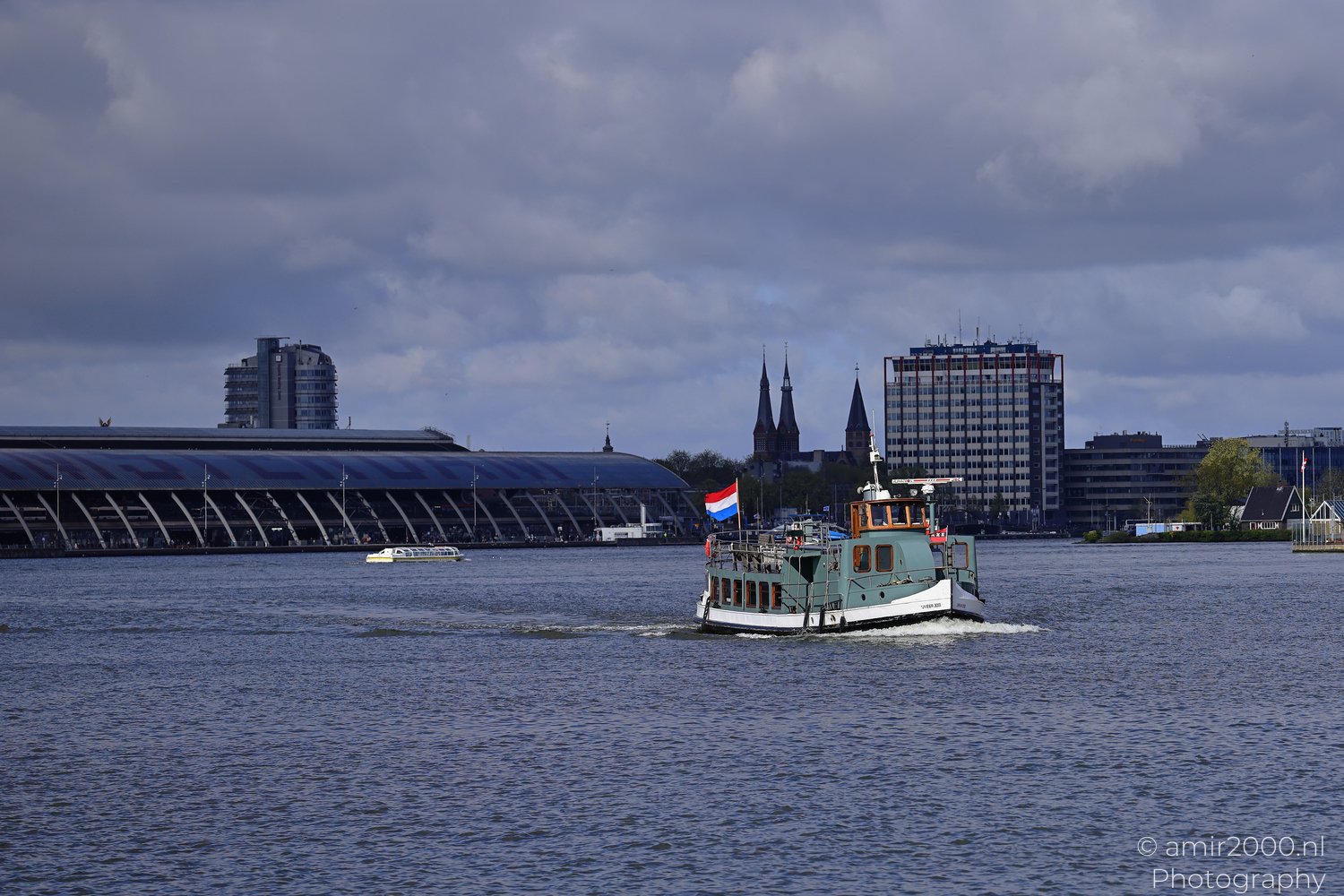 A ferry boat glides across a wide river under an overcast sky, with buildings and flags visible along the shore.