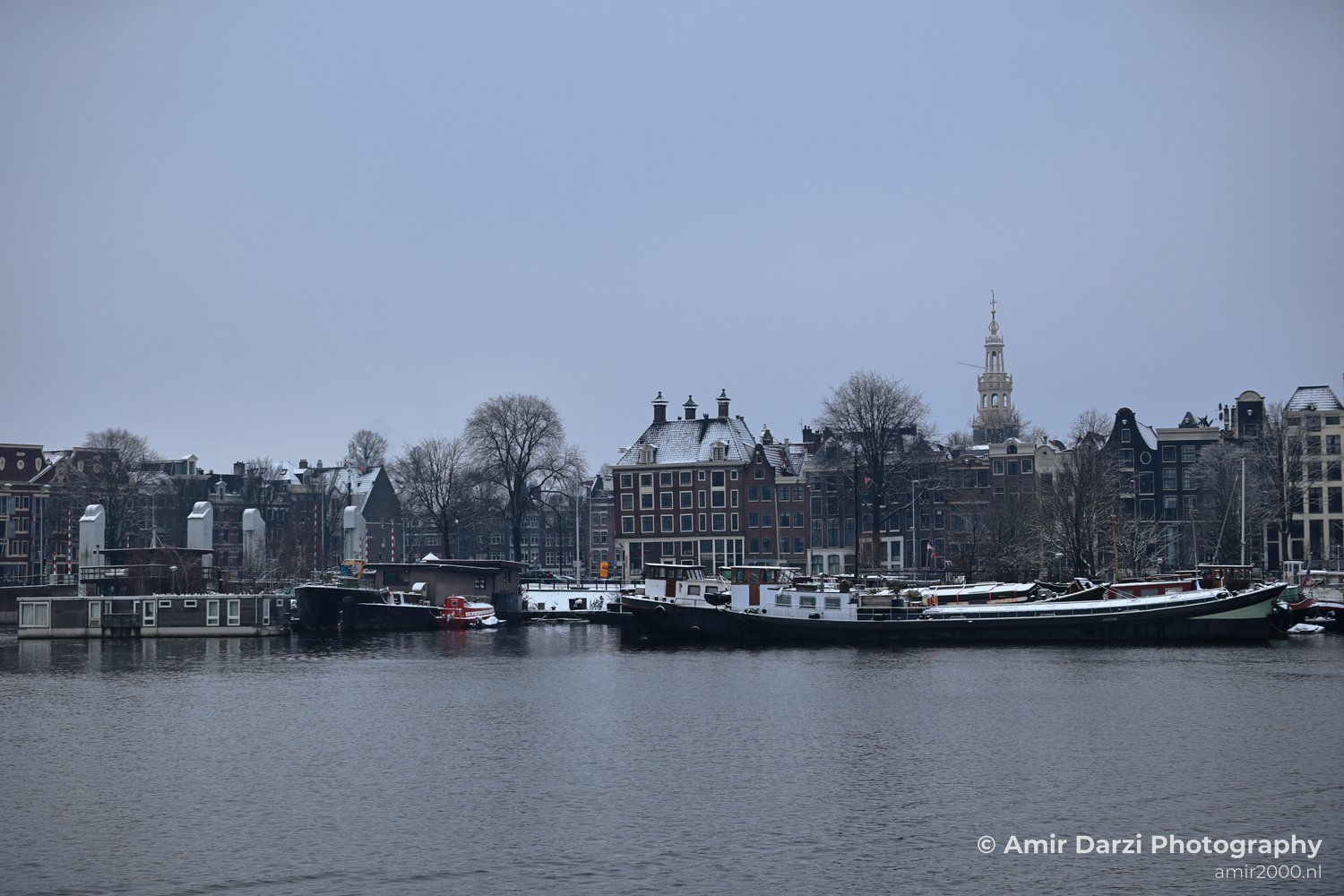 Snowy day along Prins Hendrikkade featuring boats docked near historic architecture, Amsterdam Netherlands.