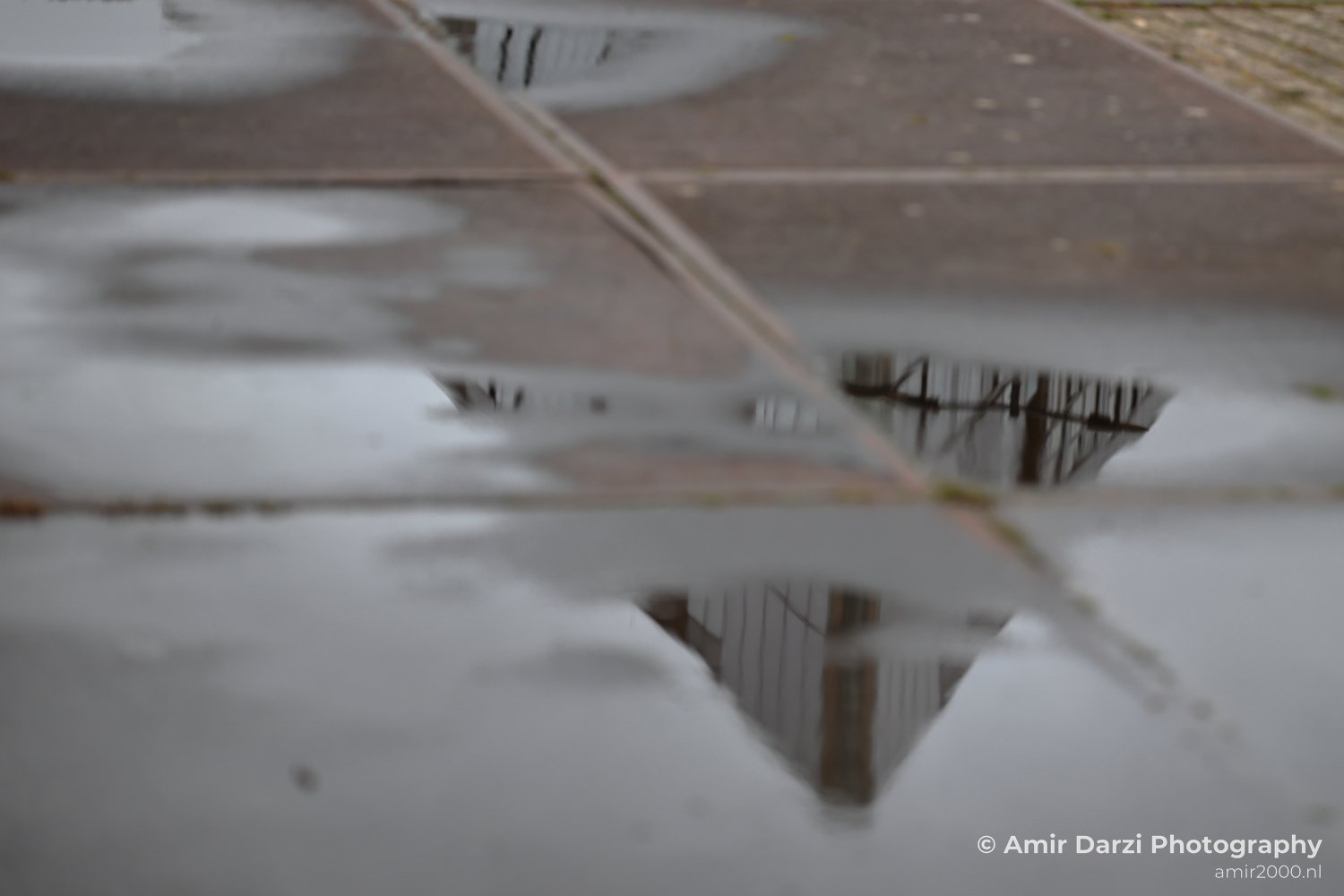 A reflection of an architectural structure and surrounding environment seen in water accumulated from recent rainfall.