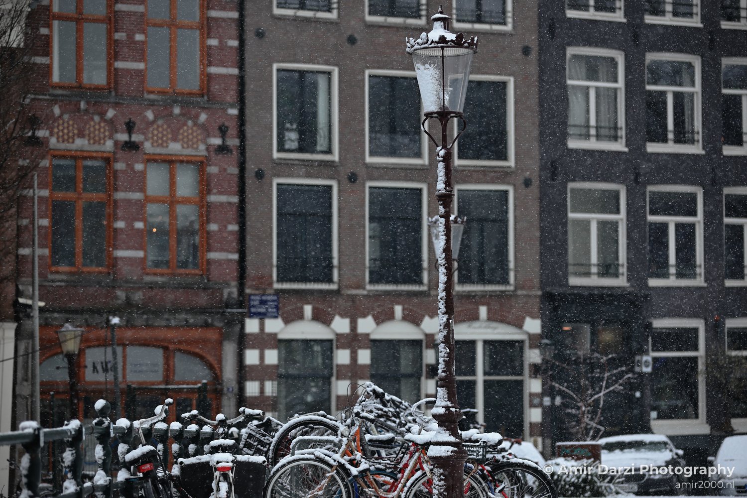 Snowy lamp and parked bikes near brick buildings in Amsterdam's Canal District.