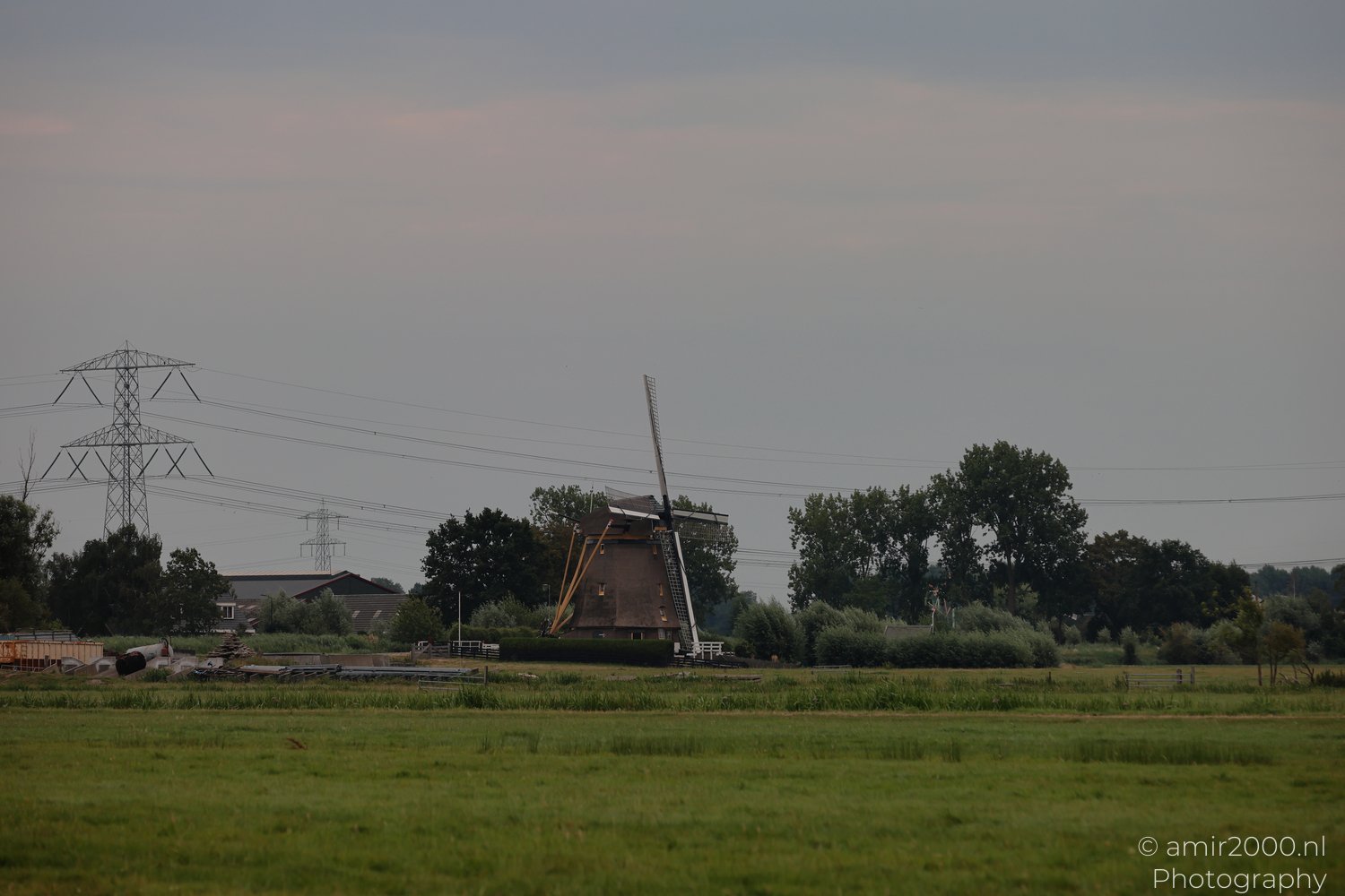 A rustic windmill is nestled in a rural landscape with lush greenery and distant power lines in Amsterdam, Netherlands.